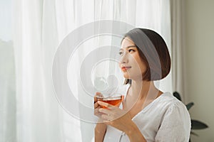 Portrait of a satisfied woman thinking looking at side holding a tea mug beside a window at home