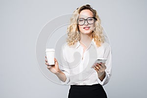 Portrait of a satisfied young business woman using mobile phone while holding cup of coffee to go over white background