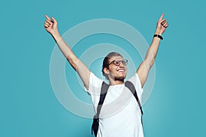 Portrait of a satisfied young man wearing white tshirt and bagpack celebrating success, isolated over blue background.
