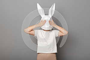 Portrait of scared anonymous woman wearing white t shirt and paper rabbit mask standing covering her eyes with palms to not see