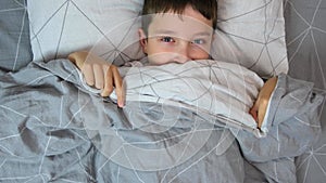 Portrait of a scared boy in bed getting out from under the cover and covering his face with blanket, children kid fears concept