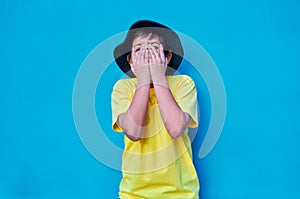 Portrait of scared boy with hands covering his face with yellow t-shirt, on blue background. Copy space