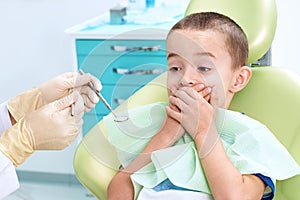 Portrait of a scared child in a dental chair. The boy covers his mouth with his hands, afraid of being examined by a