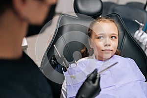 Portrait of scared cute little girl sitting in dental chair and looking with fear to unrecognizable female dentist