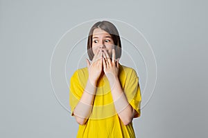 Portrait of a scared teen girl in yellow casual t shirt, looking away with wide opened eyes, covering mouth her hands, stand over