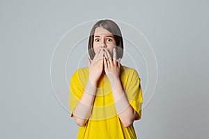 Portrait of a scared teen girl in yellow casual t shirt, looking at camera with wide opened eyes, covering mouth her hands, stand
