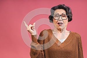 Portrait of schocked amazed surprised beautiful senior mature aged woman demonstrating copy space isolated over pink background, photo