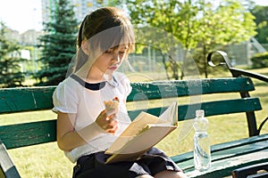 Portrait of schoolgirl 7 years old on a bench reading book, eating ice cream. Background city park