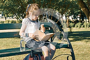 Portrait of schoolgirl 7 years old on a bench reading book, eating ice cream. Background city park