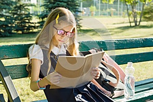 Portrait of schoolgirl primary school with backpack on a bench reading book, background school yard