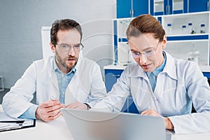 portrait of scientists in lab coats and eyeglasses working on laptop together at workplace