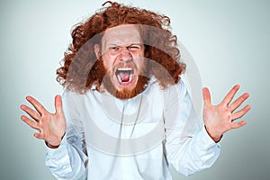 Portrait of screaming young man with long red hair and shocked facial expression on gray background