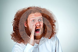 Portrait of screaming young man with long red hair and shocked facial expression on gray background