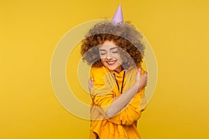 Portrait of selfish curly-haired hipster girl with funny cone hat embracing herself and smiling with pleasure