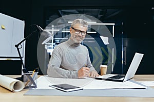 Portrait of senior architect, gray haired designer smiling and looking at camera, man mock up plan use laptop and tablet