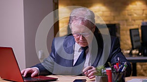 Portrait of senior businessman in formal costume working with laptop and smartphone simultaneously in office.