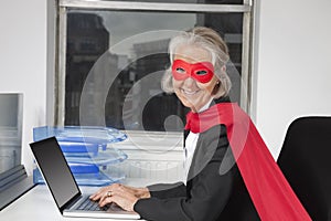 Portrait of senior businesswoman in superhero costume using laptop at office desk