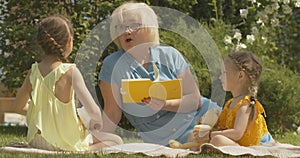 Portrait of senior Caucasian woman in eyeglasses reading book for cute little girls outdoors. Happy grandmother spending