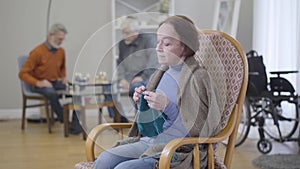 Portrait of senior Caucasian woman knitting and looking back at men playing chess at the background. Lifestyle of