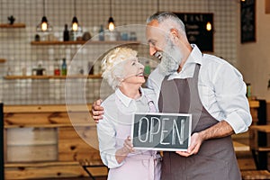 portrait of senior coffee shop owners in aprons holding open chalkboard together