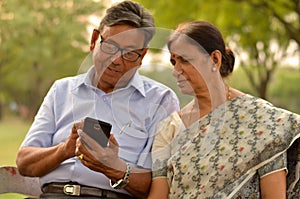 Portrait of senior couple sitting in park bench and looking at their smart phone and laughing in New Delhi, India with focus on