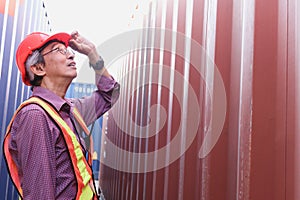 Portrait of senior elderly Asian worker engineer wearing safety vest and helmet, standing between red and blue containers