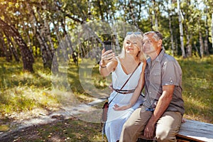 Portrait of senior family couple taking selfie using smartphone in summer forest. Elderly people rest sitting on bench