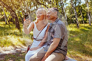 Portrait of senior family couple taking selfie using smartphone in summer forest. Elderly people rest sitting on bench