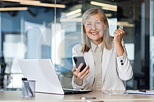 Portrait of a senior gray-haired businesswoman working in the office at a laptop, holding a phone in her hands, happy