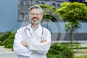 Portrait of senior gray-haired doctor, mature man in white medical coat with crossed arms and stethoscope smiling and