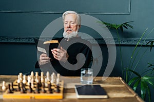 Portrait of senior gray-haired older male reading paper book at home sitting at wooden table with chess board, relax and