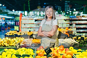 Portrait of a senior gray-haired woman of a supermarket worker, worker spreads fruit with crossed arms smiles and looks at camera