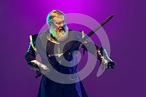 Portrait of senior greybearded man, brave and brutal medieval warrior or knight in armor with sword isolated over purple