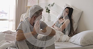 Portrait of senior handsome Caucasian man sitting on bed edge and looking back at wife lying at the background. Mature