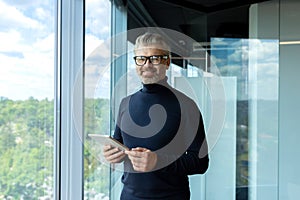 Portrait of a senior handsome gray-haired businessman in glasses standing in the office of a skyscraper by the window