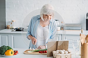 portrait of senior lady looking for recipe in cookery book while cooking dinner at counter photo