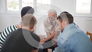 Portrait of senior male psychologist and diverse group of men and women sitting in circle and holding crosswise hands