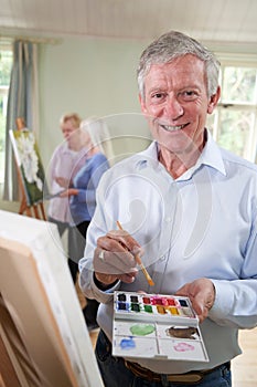 Portrait Of Senior Man Attending Painting Class With Teacher In Background