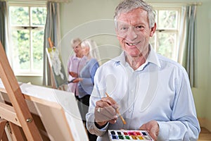 Portrait Of Senior Man Attending Painting Class With Teacher In Background