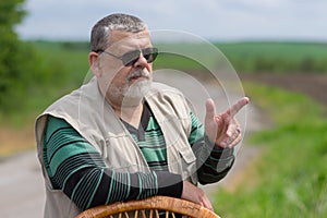 Portrait of senior man in black sunglasses leant on a wicker chair back and gesticulating