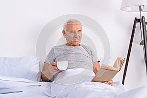 portrait of senior man with cup of coffee reading book in bed