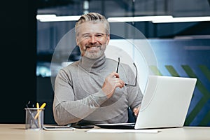 Portrait of senior man inside office, mature businessman with beard smiling and looking at camera, boss working at desk