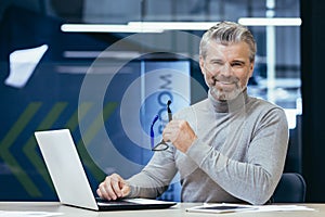 Portrait of senior man inside office, mature businessman with beard smiling and looking at camera, boss working at desk