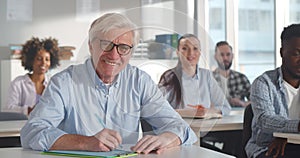 Portrait of senior man student writing at desk and smiling at camera in classroom