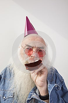 Portrait of senior man wearing party hat and red glasses while blowing horn against gray background