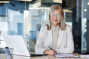 Portrait of senior mature female financier at workplace, gray haired businesswoman smiling and looking at camera doing