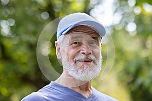 Portrait of senior mature gray-haired man in park, pensioner in sports cap smiling and looking at camera close-up among