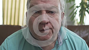 Portrait of senior mature old man with blue eyes sitting on the sofa at home eating chips close up. Grandfather looking