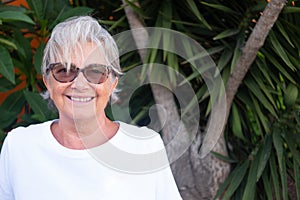 Portrait of senior pretty woman with gray hair. Sitting outdoor in the garden. Background of plants and leafs. Smiling and