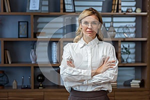 Portrait of senior serious female boss in classic office in the evening, mature business woman with crossed arms looking